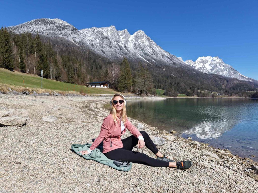 Hintersteiner See im Frühling, Eine Frau sitzt am Ufer eines klaren Sees, umgeben von schneebedeckten Bergen. Sie trägt Sonnenbrillen und lächelt.