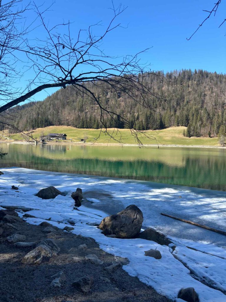 Hintersteiner See im Frühling, Ein ruhiger See mit schneebedecktem Ufer, umgeben von Bäumen und einem klaren blauen Himmel.