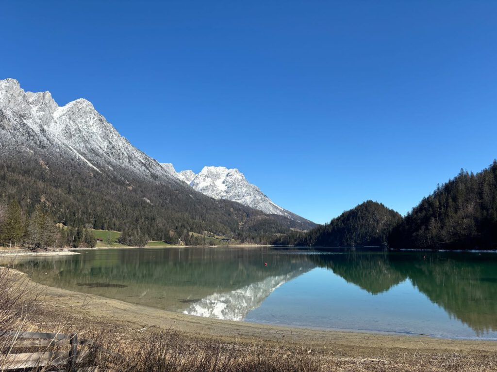 Hintersteiner See im Frühling, Ruhiger Bergsee mit klarer Reflexion der Berge und blauem Himmel, umgeben von Wäldern und schneebedeckten Gipfeln.