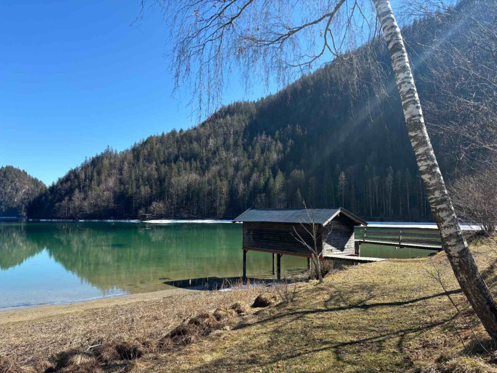 Hintersteiner See im Frühling, Ein ruhiger See mit einem Holzhaus am Ufer, umgeben von Bergen und Bäumen unter klarem blauen Himmel.