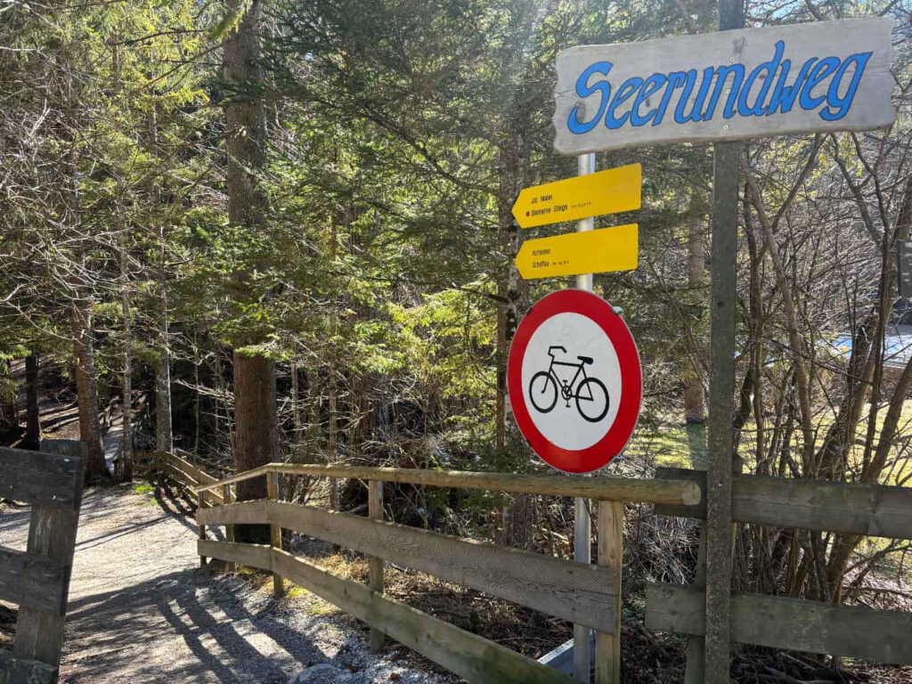 Hintersteiner See im Frühling, Schild des Seerundweges mit Hinweis auf Radfahrverbot und Wegbeschreibung in einem Waldgebiet.