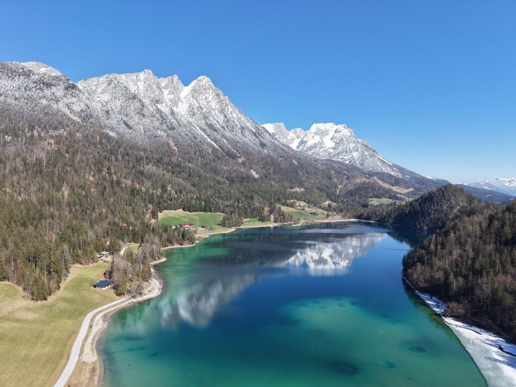 Hintersteiner See im Frühling, Eine panoramaartige Ansicht eines klaren Sees, umgeben von bewaldeten Hügeln und majestätischen Bergen im Hintergrund, bei strahlend blauem Himmel.