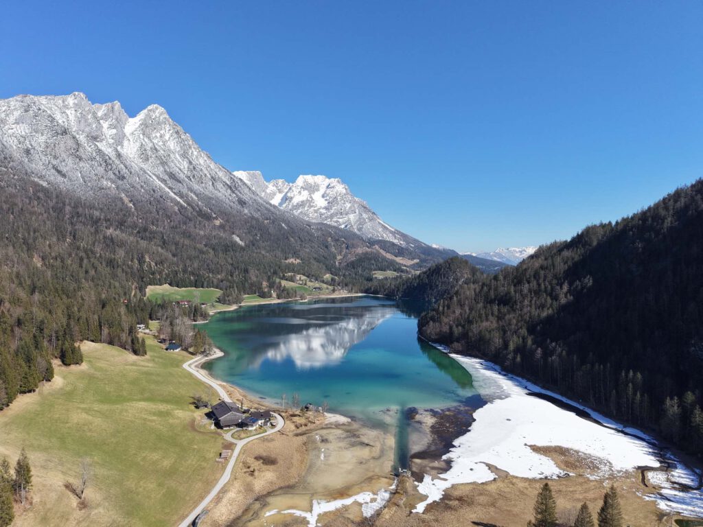 Hintersteiner See im Frühling, Luftaufnahme eines klaren Sees, umgeben von schneebedeckten Bergen und grünen Wiesen im Frühling.